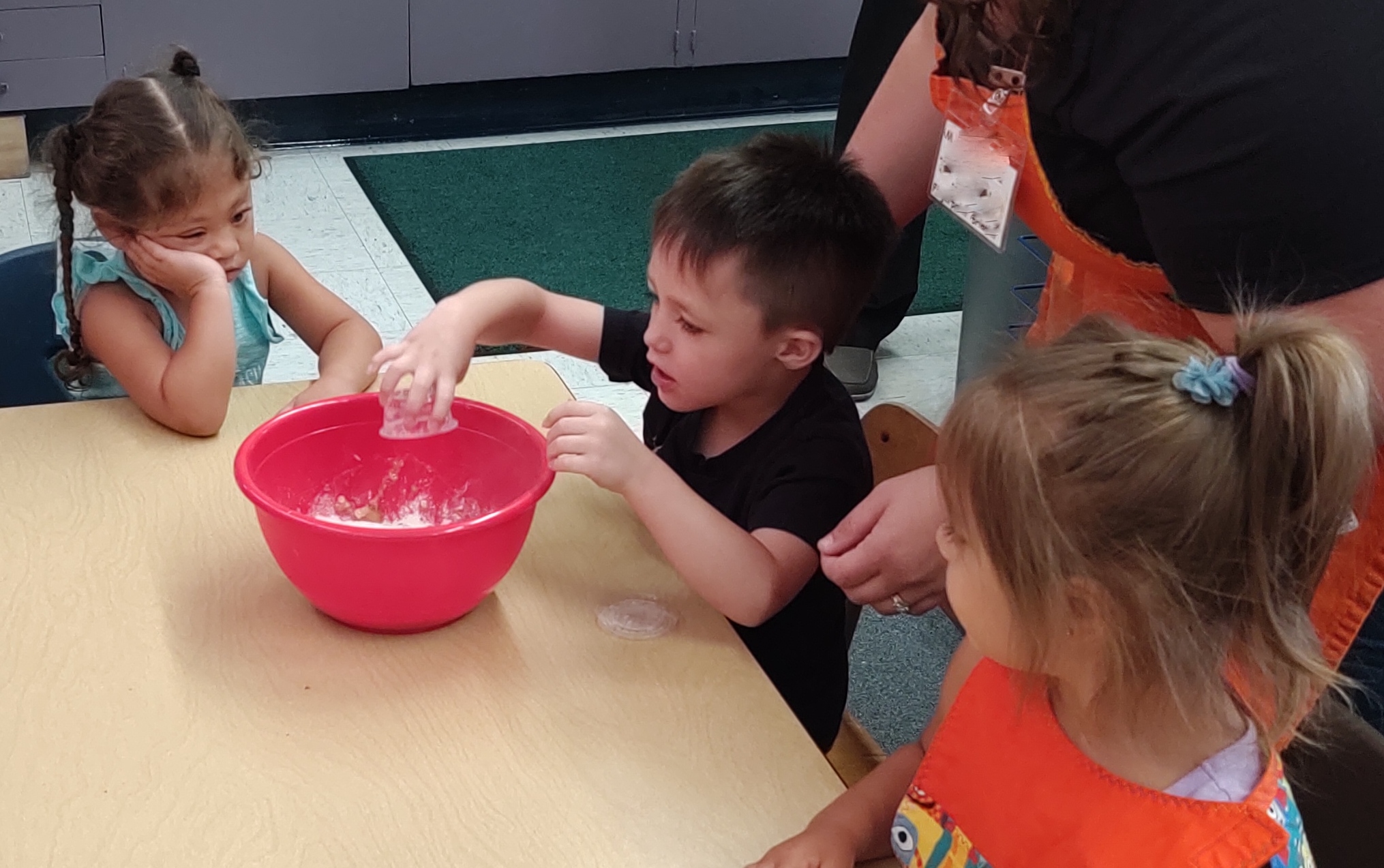 Two girls watching a boy add ingredients to a red bowl as they make muffins.